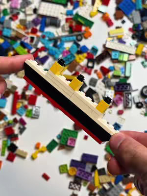 A miniature Lego Titanic is held between two hands. The ship is white with black and red accents, and yellow smokestacks. It is surrounded by a scattered collection of colorful Lego bricks on a white surface.