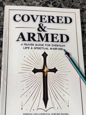 The image showcases the cover of a book titled 'COVERED & ARMED'. The book's design is mostly white with black and gold text and illustrations. The focus is on a black sword-shaped cross centered on the cover, with golden rays emanating from behind it. A pen is positioned diagonally across the top right corner. Overall, the composition is clean and spiritual.