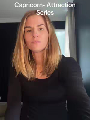 A woman with light brown hair looks directly at the camera with a neutral expression. The top half of her upper body is visible, wearing a black long-sleeved shirt. Above her head is a text overlay that says "Capricorn- Attraction Series". The backdrop is a blurry and neutral color palette.