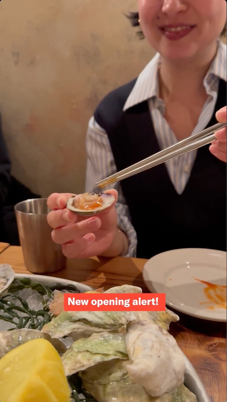 The image shows a woman with short dark hair smiling while holding an opened oyster with reddish sauce in one hand and metal chopsticks in the other. She is wearing a black vest over a striped shirt, and a glimpse of a partially visible stainless steel cup and plate appear in the background. Oysters on ice are in the lower part, the scene is well-lit.