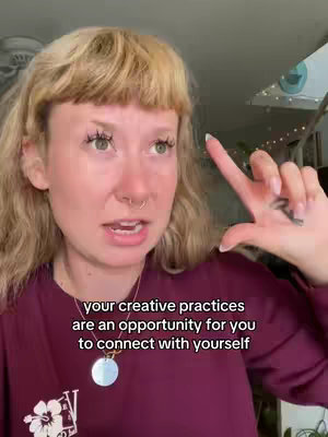 A woman with blonde hair and a nose piercing is holding up her right hand with the index finger extended, pointing upwards. She's wearing a maroon shirt and a necklace. Text overlayed at the bottom says 'your creative practices are an opportunity for you to connect with yourself'.