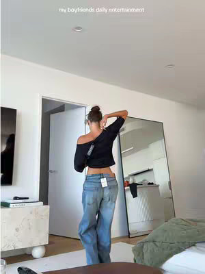 A person, viewed from the waist up and from the back, stands in a bright room, turned slightly to the right. They wear a black off-the-shoulder top and light blue jeans. The person is styled with hair pulled up and a handbag on their shoulder. Behind them, a full-length mirror reflects part of a modern white and light-colored interior. Some text is added overhead.