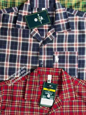 The image shows a close-up view of two plaid shirts, one navy and one red. The navy shirt is on top with a green tag, while a red shirt rests underneath with a yellow tag. The shirts fill most of the frame, showing the fabric's pattern, collar, pocket, and tags.