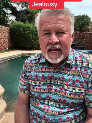 A middle-aged man with gray hair and a beard stands outdoors near a pool, wearing a colorful patterned shirt. Text overlay in a red banner at the top reads 'Jealousy'.