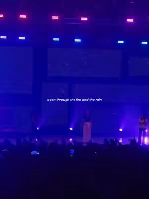A live performance on a stage, with the audience in the foreground. The lighting is primarily blue and purple, with spotlights on the performers. Text overlayed at the center reads 'been through the fire and the rain.'  The overall composition conveys excitement of a concert or worship event.