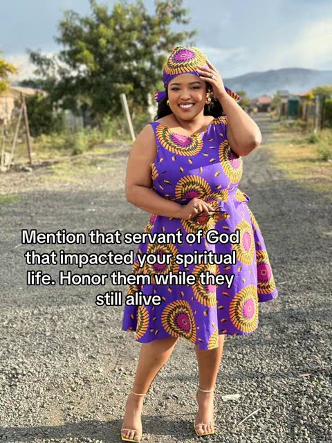 A medium shot features a smiling woman in a colorful outfit against a blurred outdoor background. The woman is the focal point, holding her hair, wearing a headscarf and dress with a repeating circular pattern in purple, yellow, and orange. The background is a mix of greenery, overcast sky, and a gravel path. The overall composition is well-lit, with a focus on the woman's pose and attire.