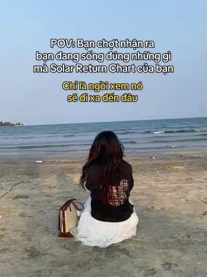 A woman is sitting on a sandy beach, facing the ocean. She is wearing a black top and white skirt, and a handbag is next to her. Text overlays in Vietnamese. The sky is partly cloudy.