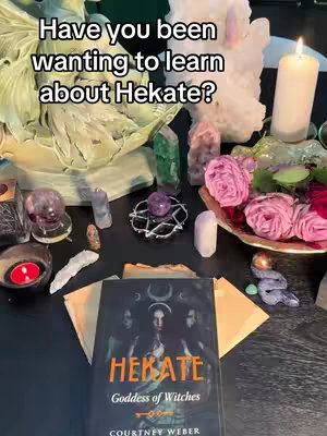 The image shows a dark wooden surface with a collection of items related to spirituality and witchcraft. In the center, a book titled 'HEKATE Goddess of Witches' is displayed, surrounded by crystals of various shapes and colors (clear, amethyst, green). A lit candle is prominent on the right side, with pink roses and a dish containing more crystals adding to the mystical aesthetic. The overall composition suggests themes of spirituality, magic, and knowledge.