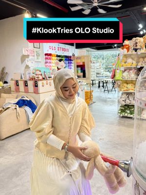 A person is seated in a DIY studio, holding a plush bunny. They are wearing a beige headscarf and a cream-colored outfit, set against a background of craft supplies and other plush toys displayed on shelves. Above the subject is a text overlay with a call-to-action (#KlookTries OLO Studio) in a black, red and white rectangular box. The overall scene has a warm, inviting, and creative atmosphere. There's a claw machine on the right side.