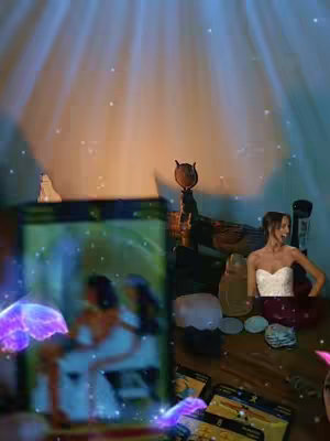 The image shows a tarot card reading scene. A woman in a white dress stands near a table with tarot cards and decorative objects.  Overlayed on the image are soft, blue light beams, stars, and swirling purple effects. A central tarot card is blurred, but shows two blurred figures.