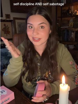 A young woman with dark hair and a light green hoodie sits at a table. The image is dimly lit, with a lit candle to the right. In the foreground, she gestures with one hand and appears to be speaking. On the table are some items, including cards. Text overlays on the image read self discipline AND self sabotage.