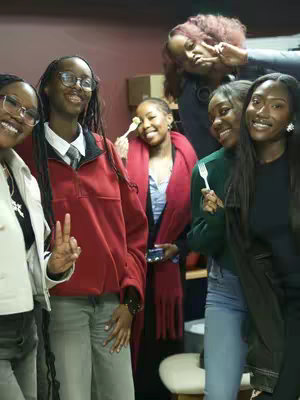 A group photo featuring several young, Black women smiling and posing. They are in a warmly lit room. Some are holding forks, and most are smiling at the camera, with a variety of outfits and accessories.