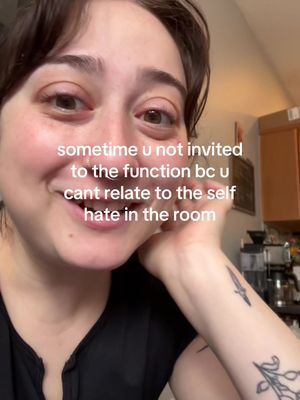 A selfie featuring a person with short, dark hair smiling and looking towards the camera. The image is a close-up, showing the person's face and part of their upper body. Text overlayed on the image reads "sometime u not invited to the function bc u cant relate to the self hate in the room". Behind the person, there are glimpses of a kitchen setting, including a countertop and appliances. The person wears a black top and has tattoos on their arm.