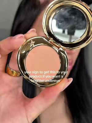 A close-up shot features a woman's hand holding a gold compact, with the open lid revealing a peachy-colored concealer. The woman's face is partially visible in the background, with black hair. Text is overlaid in the frame with instructions.