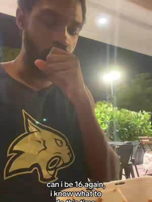 A selfie-style video cover image shows a young man in a dark shirt, with a golden cat logo, looking pensive. The backdrop is a blurry outdoor scene with trees and a street lamp. He has a hand near his mouth, suggesting thought or contemplation. A text overlay appears at the bottom.