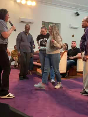 A group of people are standing in what appears to be a church setting, with a purple carpet. The focal point is on a woman holding a microphone on the left, with a casual conversation going on among the group, and a woman in jeans the center.