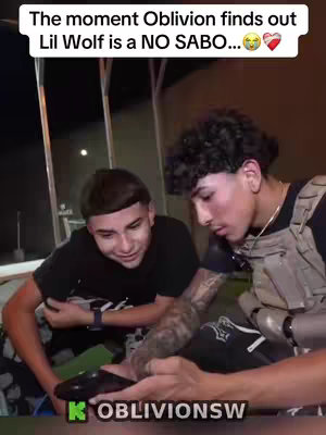 Two young men are seated and interacting. The image is framed with text at the top. One man is looking on with concern while the other holds a black object. A logo in green and white text is at the bottom.