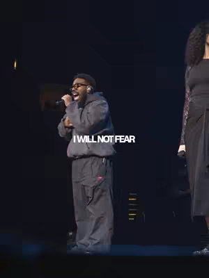 The image features a man on a stage, singing into a microphone. The man has glasses and is wearing a gray hoodie and pants. Behind him to the right a woman is standing. Below the man, the added text says 'I WILL NOT FEAR'. The background is dark, and the lighting seems part of a concert or performance.