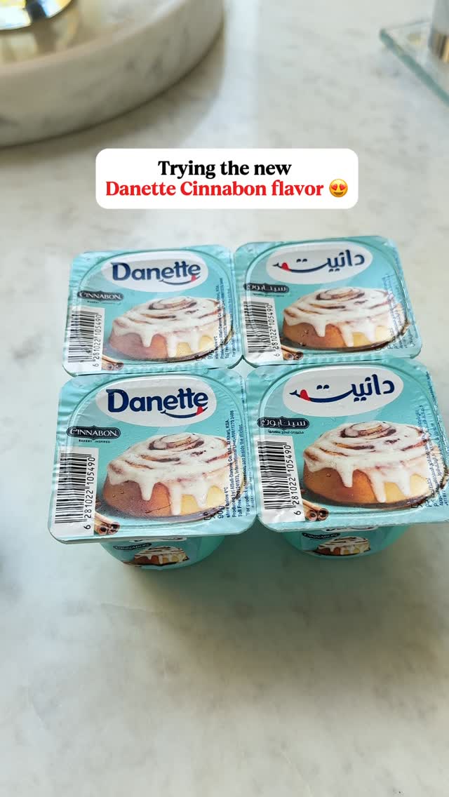 The image presents four individual blue Danette custard cups arranged in a square formation. Each cup displays a picture of a cinnamon roll with white frosting. The text overlay at the top in a white rectangular box reads "Trying the new Danette Cinnabon flavor".  The background is a light surface. The overall composition is clean and focused on product