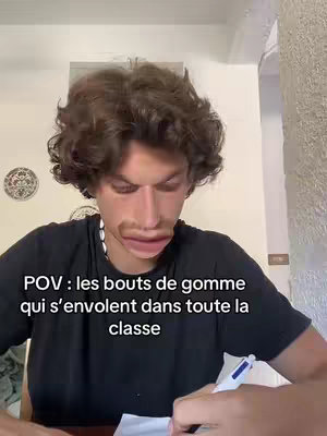 A young man is shown in a selfie-style shot, seated at a desk with the top part of a white wall visible in the background. He is writing on paper with a pen in his right hand. Overlaid is a filter that deforms his face and highlights his mouth. Text at the bottom of the frame reads "POV : les bouts de gomme qui s'envolent dans toute la classe".