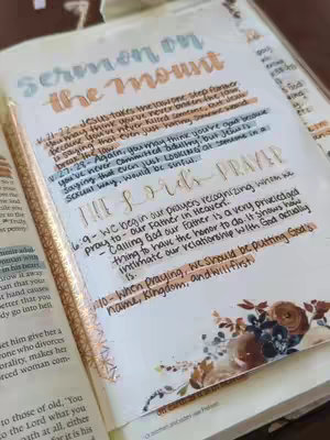 A close-up shot of an open Bible with handwritten notes and highlighting, floral decorations in the bottom right corner. The page is filled with annotations and the words \"Sermon on the Mount\" are visible, indicating a passage of scripture.