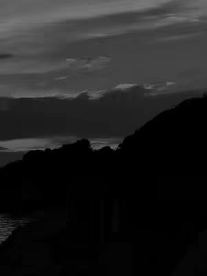 A black and white landscape photo featuring a silhouetted shoreline and a dark mountain against a cloudy sky.