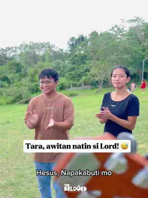 A medium shot of a green outdoor setting. Two young people are standing facing the camera, clapping. The sky is overcast. Text overlays are present, a stylized logo is at the bottom.