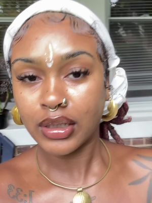 A close-up shot of a dark-skinned woman with a white headscarf and gold earrings. She has a septum piercing, a gold necklace, and a tattoo partially visible on her chest. She has makeup on and looks directly at the camera in a casual setting.