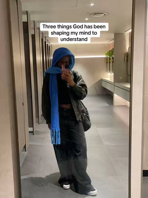 A woman is captured in a full-length mirror selfie within a restroom. She's wearing a blue hooded scarf and a matching scarf draped around her neck, a black top with a cropped fit, high-waisted black pants, a black jacket, and sneakers. She holds her phone up to the mirror to take the photo. The background shows the restroom with stalls and sinks.