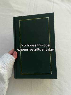 A hand holding a closed, dark green hardcover book. The book has a gold border around the cover. The background is a light-colored fabric.