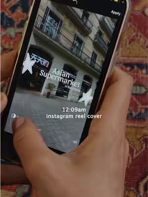 A person is holding a phone displaying a screenshot of an Instagram Reel while inside the app. The phone shows a business exterior during the day. The video is framed hands in frame, the image inside shows a building, text overlays include "Asian Supermarket" and time indicators