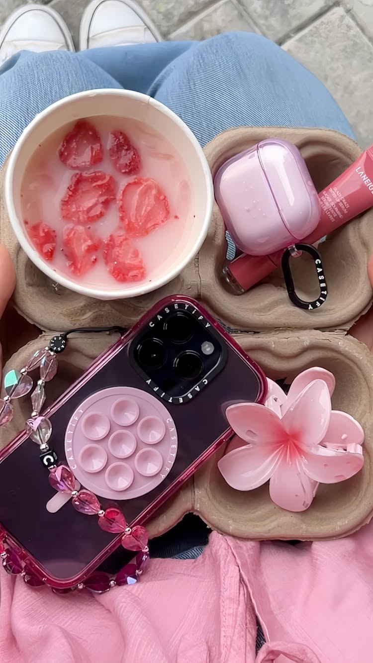 A top-down shot showing a collection of items, mostly pink, arranged in a wooden tray. The items include a cup with strawberry slices floating in a pink liquid, a phone case with a pop socket, an airpods case, lip gloss, a hair clip, and a beaded phone strap. The arrangement implies a curated, aesthetic presentation, with a focus on pastel colors and modern accessories.