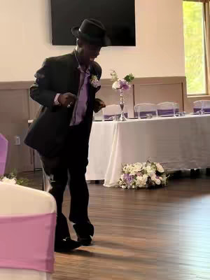 An older Black man in a suit and hat dances in what appears to be a wedding reception. He is angled slightly to the right, with his right leg forward in a dancing pose. The background suggests a formal setting, with tables and decorations.