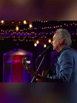 A live performance cover image. A man with light skin, gray hair on the sides, and a blue jacket is seen on stage, holding an acoustic guitar and singing into a microphone. The background is dimly lit with string lights creating an out-of-focus, warm glow and hints of red and purple lights as well.