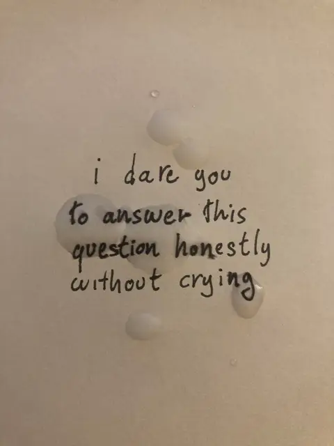 A close-up image on a white background. In the center, it features handwritten text in black ink that reads: 'i dare you to answer this question honestly without crying'.  Several irregular, light-colored blotches resembling water droplets or foam surround the text, placed somewhat randomly.