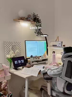 A top-down shot of a cozy study space. A desk holds a computer and a tablet showing a clock, papers, and stationery with a person sitting in a chair to the right. Behind the desk, on the wall is a display and a shelf adorned with a light and plant.