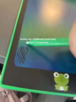 The image shows a close-up view of an electronic drawing pad with a green border. The pad is black with a digital drawing surface. A green stylus is positioned near the screen. A small, cartoon-like green frog sticker is on the bottom right corner. The words 'today my childhood goal was in fact completed' are overlaid on the screen in white text.