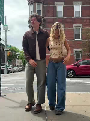 A couple is walking side by side on a sidewalk. The woman is on the right and is wearing a polka dot top and jeans, the man is on the left, the image is set in front of brick buildings.