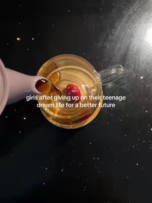 An overhead shot captures a glass apparatus with liquid and a small red item inside, possibly cherries. A hand with dark nail polish is partially visible, seemingly pouring from a pink container. The background is a glossy black surface speckled with small, bright dots. Text overlay reads "girls after giving up on their teenage dream life for a better future".