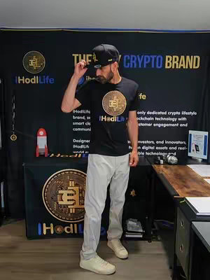 A man in front of a backdrop that says "THE CRYPTO BRAND", with a black shirt that also has crypto branding. The person is wearing a black cap, white pants and white shoes. He is touching his hat in a casual way in the middle of the shot.