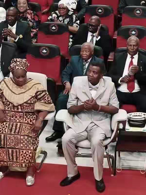The image captures a room filled with people, likely at a religious event or conference. Several individuals are seated. The focus is primarily on Pastor E.A Adeboye who is sitting and has a composed expression. The attire of the individuals suggests a formal or religious setting. The background consists of other people. The color scheme is fairly neutral, with a focus on skin tones and the dark tones of the seating and clothing, creating a straightforward and formal environment.