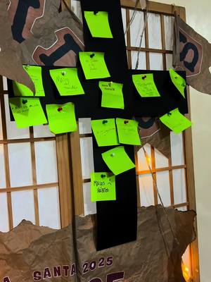 The cover image pictures a wooden structure resembling a large cross, with several small neon green sticky notes attached to it. Some notes have handwritten messages, pinned with red thumbtacks. The cross is set against a backdrop of a window, with brown paper decorations draped around the frame, partially obscuring a view of the outdoors. The overall composition suggests a religious or spiritual theme, possibly a communal activity.