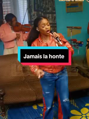 A female singer with long, dark hair is positioned center-frame, holding a microphone and singing. She's wearing a bright orange or red satin shirt over a white t-shirt, paired with blue jeans. Behind her, a person is playing the violin and a blurred background with musical equipment such as drums and a conga drum.  A text overlay that reads, 'Jamais la honte' is displayed in a black and blue rectangle.