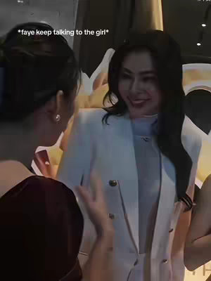 The image shows two women in a setting that appears to be an event or gathering. The woman in focus is wearing a white blazer with gold buttons and there is text overlayed on the top left corner of the image.