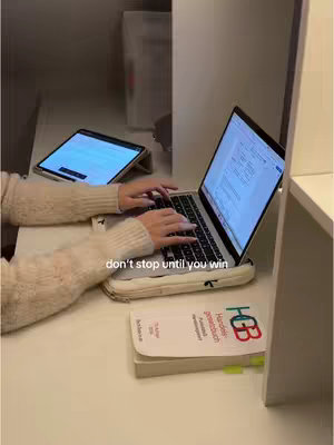 A top-down shot showing a person at a desk with a laptop and tablet open, typing. A textbook is visible, along with a motivational quote overlay.