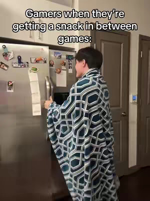 A person wrapped in a geometric patterned blanket opens a refrigerator door in a well-lit kitchen. The person is turned to the side, with the refrigerator to their left and a closed door visible in the background.