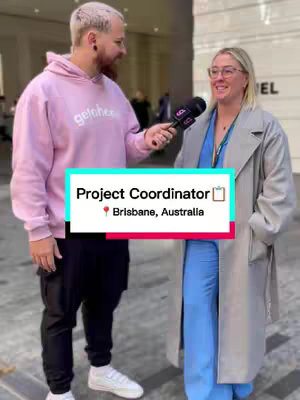 A man in a pink hoodie is interviewing a woman in a grey overcoat. Both are standing outside. A graphic overlay in the center highlights 'Project Coordinator' and 'Brisbane, Australia.'