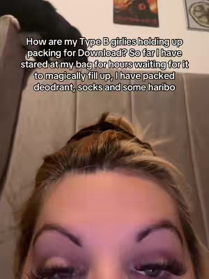 A selfie-style shot showing the face of a person. The person has light-colored hair up in a bun with some strands falling down. The person has prominent eyelashes and is wearing makeup including eyeshadow. The background is blurred and appears to be an indoor setting.
