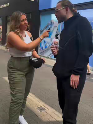 A woman with blonde hair is interviewing a man on a city street. The woman is holding a microphone and gesturing. The man wears glasses and is holding a small, rounded pink object, a gift, at his waist height. The city street is visible in the background.