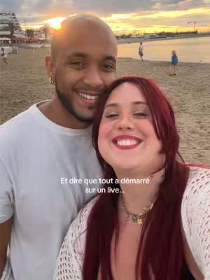 A selfie of a smiling couple on a beach at sunset. The woman has red hair and the man is bald. Both are smiling broadly and looking at the camera. The background shows a beach with people, the ocean, and a golden sunset.